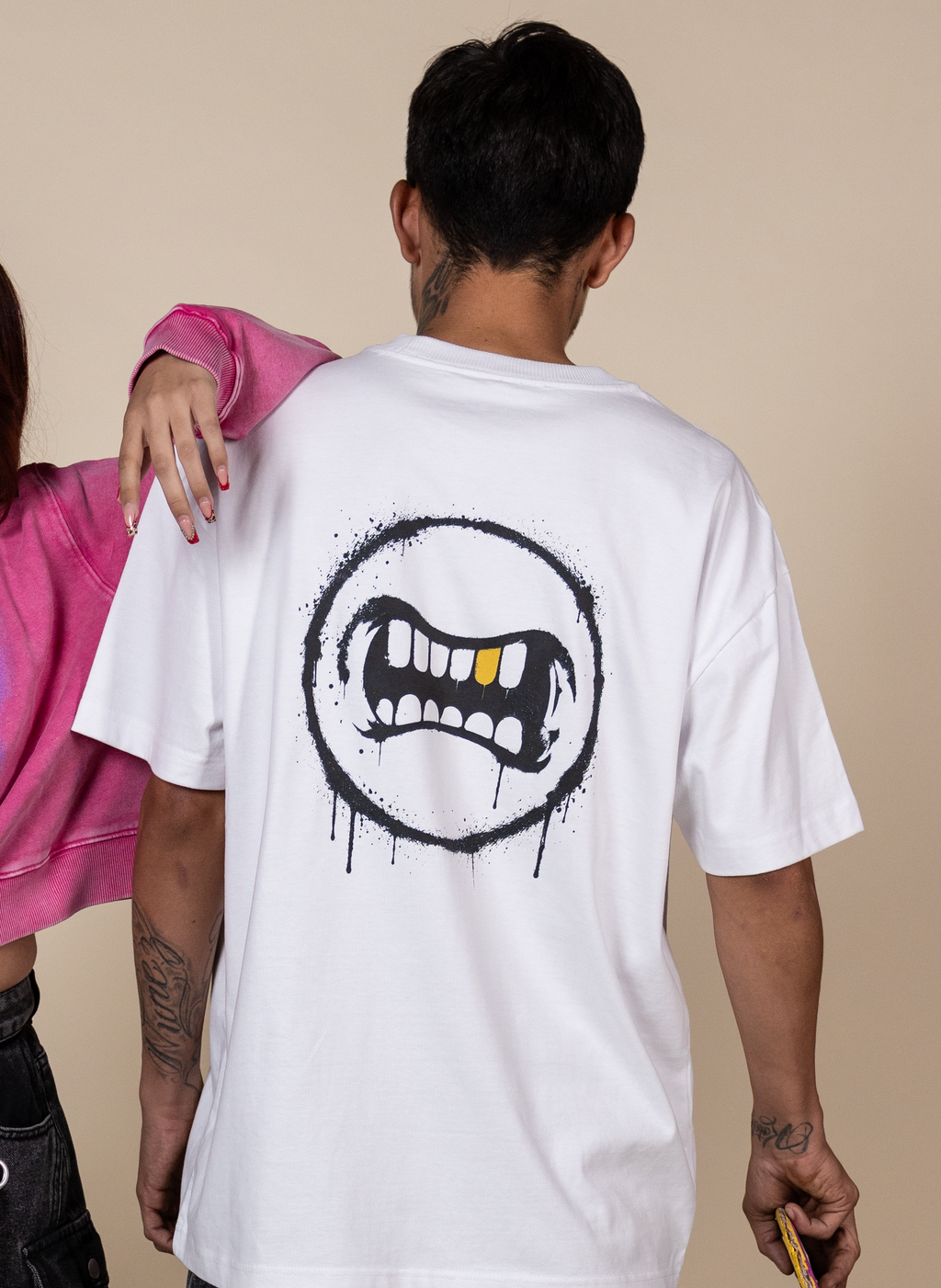 A man wearing a white oversized T-shirt from the back, featuring a black graffiti-style mouth graphic with yellow teeth accents. A woman in a pink sweatshirt is partially visible on the left, and the background is a solid beige studio wall.