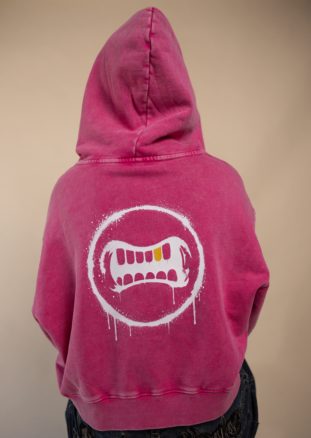 A person shown from the back wearing a bright pink hooded sweatshirt with a large white dripping mouth graphic on the back, paired with dark denim shorts, against a plain beige background.