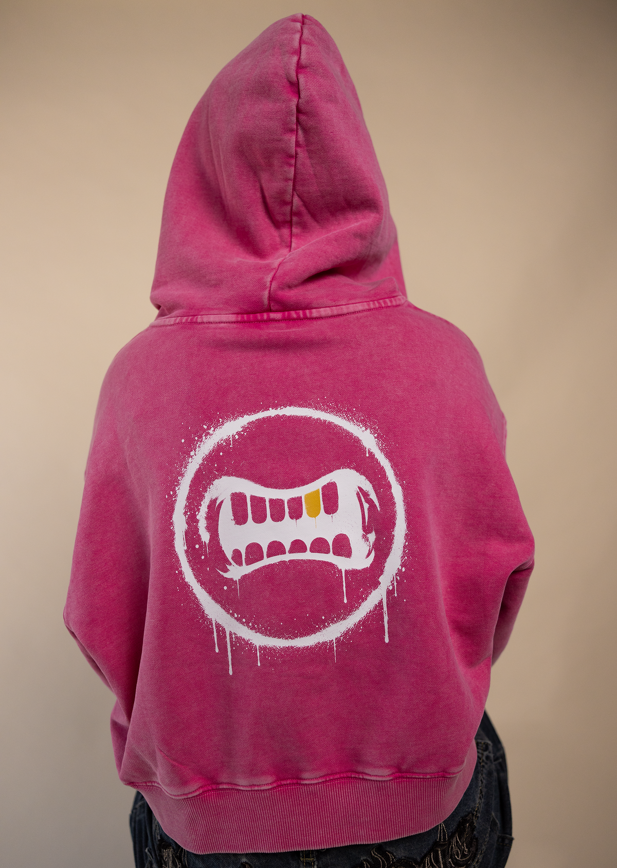 A person shown from the back wearing a bright pink hooded sweatshirt with a large white dripping mouth graphic on the back, paired with dark denim shorts, against a plain beige background.