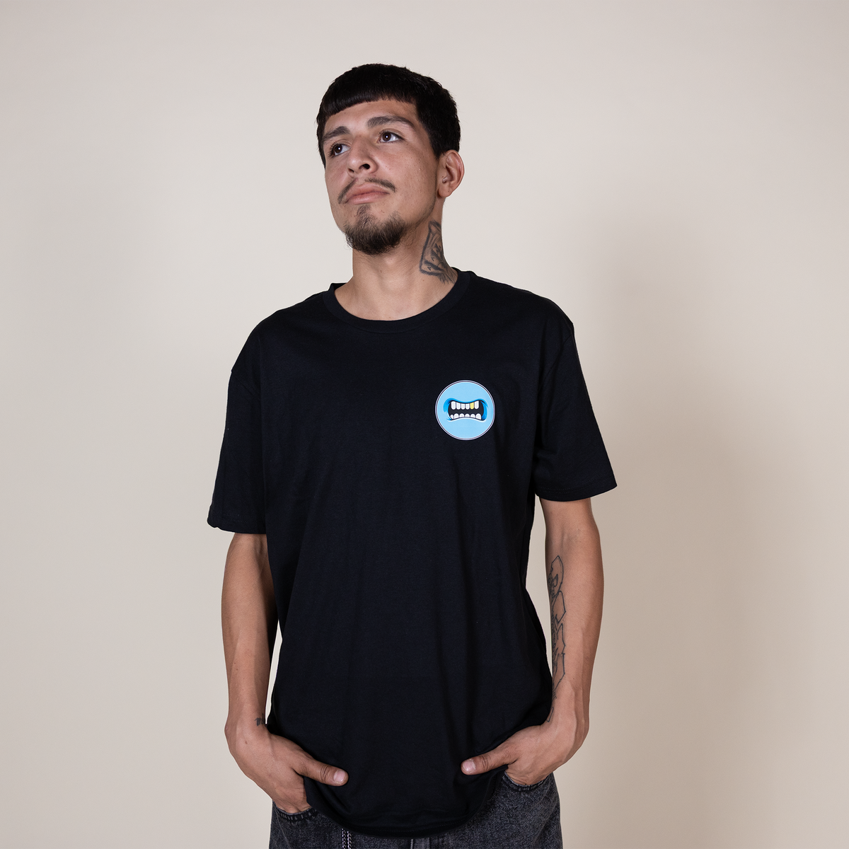 A man wearing a black crewneck t-shirt with a circular blue and white graphic on the left chest, standing against a plain beige background.