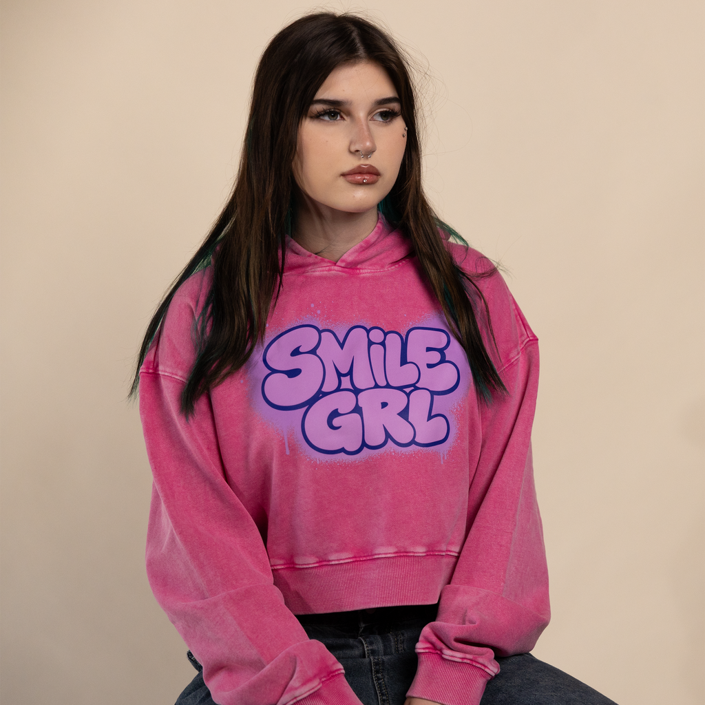 A young woman with long dark hair sits wearing a pink acid-wash cropped hoodie with 'SMILE GRL' printed in purple bubble letters on the front, paired with dark blue jeans against a solid beige background.